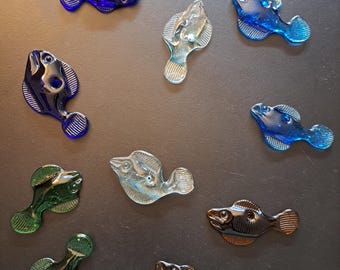 Könnte beinhalten: Eine Sammlung von Glasfischfiguren in verschiedenen Farben, darunter Blau, Grün und Klar. Die Fische haben detaillierte Merkmale und sind auf einer dunklen Oberfläche angeordnet. Die Figuren sind kleine Dekorationsartikel.