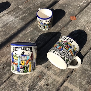 Pode incluir: Três canecas de cerâmica com acabamento azul e branco. Cada caneca apresenta uma ilustração colorida de um horizonte urbano com o nome da cidade impresso em preto. As canecas são "Jerusalém", "Belém" e "Salem".