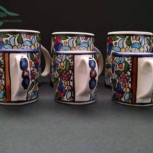 May include: A set of six ceramic mugs with a floral design. The mugs have a white base with colorful flowers, leaves, and blue accents. The handles are white with blue details, and the rims are blue. The image also shows the text "MYSTIC LAND PAINTED" in the upper left corner.
