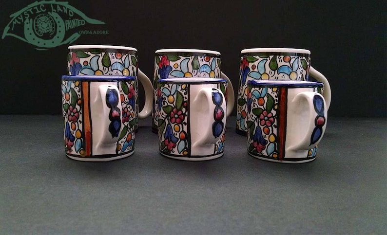 May include: A set of six ceramic mugs with a floral design. The mugs have a white base with colorful flowers, leaves, and blue accents. The handles are white with blue details, and the rims are blue. The image also shows the text "MYSTIC LAND PAINTED" in the upper left corner.
