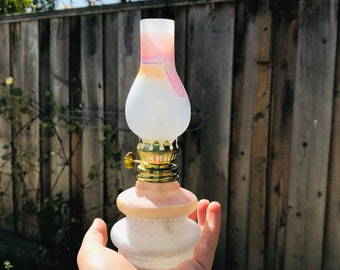 May include: A small, antique-style oil lamp with a frosted glass shade and a pink and white base. The lamp has a brass-colored metal top and a decorative design. The lamp is held in a hand against a wooden fence background.