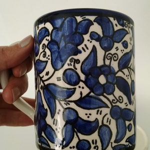 May include: A ceramic mug with a white background and a blue floral pattern. The mug has a white handle and a black rim. The floral design features blue flowers and leaves with black outlines. The mug is part of a set.