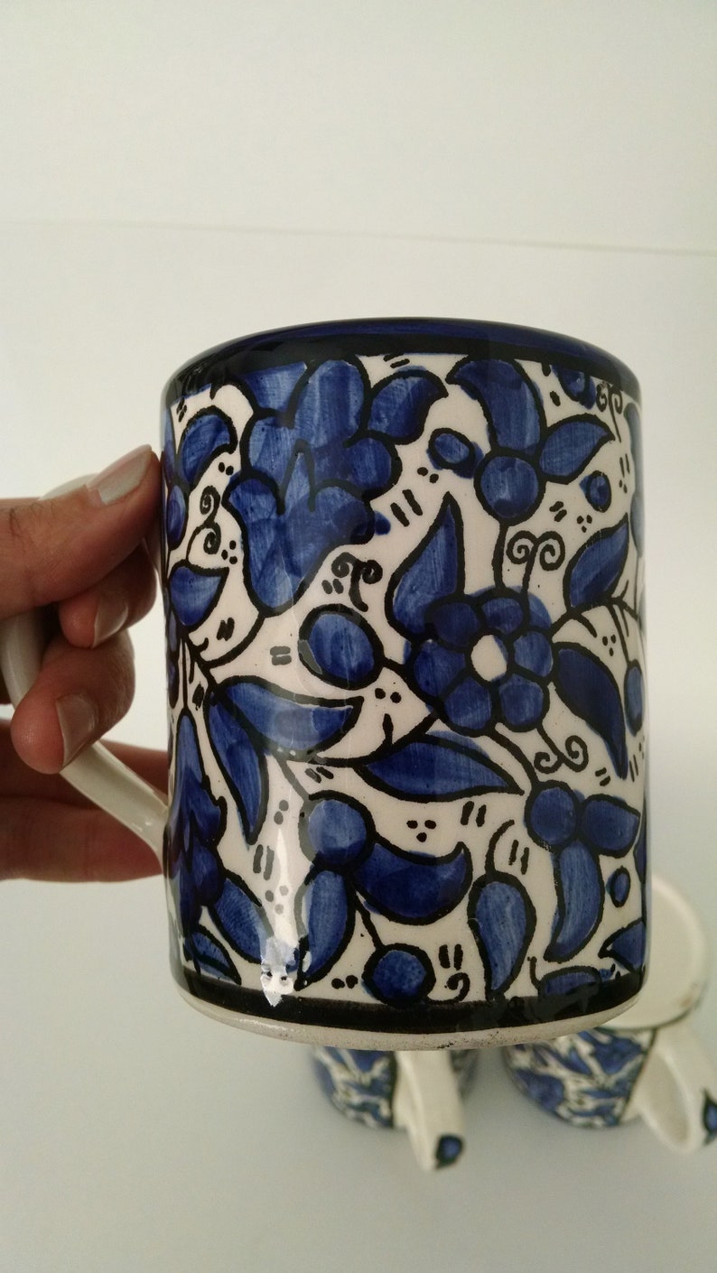 May include: A ceramic mug with a white background and a blue floral pattern. The mug has a white handle and a black rim. The floral design features blue flowers and leaves with black outlines. The mug is part of a set.