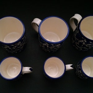 May include: Six ceramic mugs with white interiors and handles. The mugs have a blue floral pattern on the exterior and a blue trim around the rim. The mugs are arranged on a dark surface.
