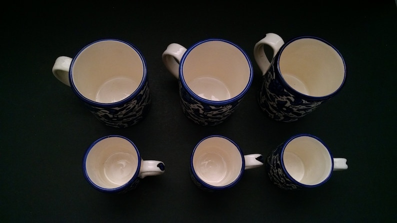 May include: Six ceramic mugs with white interiors and handles. The mugs have a blue floral pattern on the exterior and a blue trim around the rim. The mugs are arranged on a dark surface.