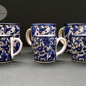 May include: A collection of six ceramic mugs with a blue floral pattern on a white background. Each mug has a white handle and a dark blue rim. The mugs vary slightly in size and are arranged on a dark surface.