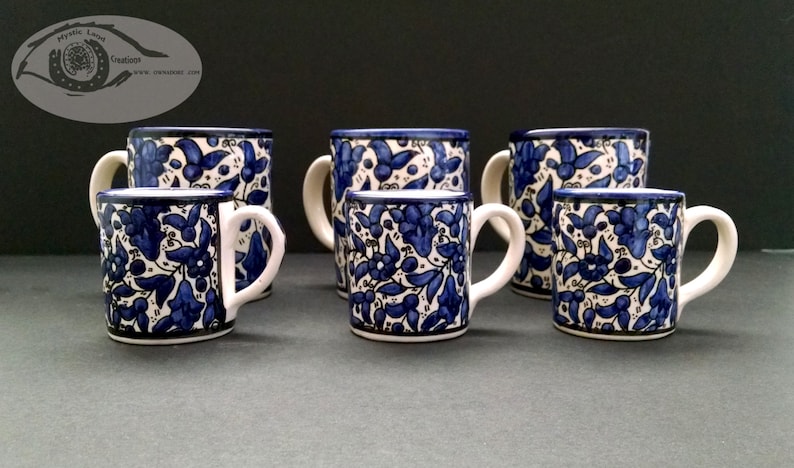 May include: A collection of six ceramic mugs with a blue floral pattern on a white background. Each mug has a white handle and a dark blue rim. The mugs vary slightly in size and are arranged on a dark surface.