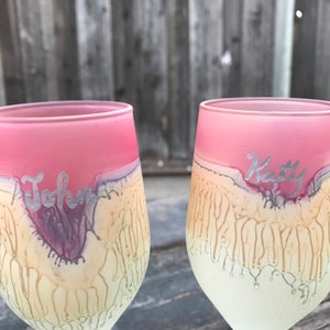 Może przedstawiać: Dwa spersonalizowane kieliszki do wina z różową obwódką i kremową podstawą. Kieliszki mają srebrny napis "John" i "Katy". Projekt zawiera efekt kapania w odcieniach kremu i fioletu.