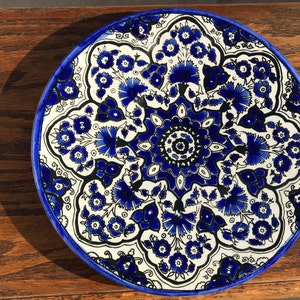 May include: A round, decorative ceramic plate with a blue floral design. The plate features a white background with intricate blue flower and leaf patterns, outlined in black. The rim is a solid blue.