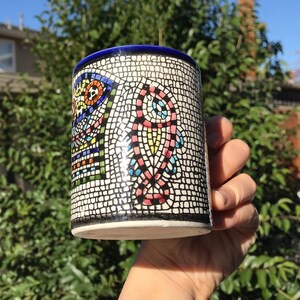 May include: A cylindrical ceramic mug with a mosaic design. The mug features a white background with a black grid pattern, and colorful mosaic art in shades of red, blue, yellow, and green. The rim is blue.