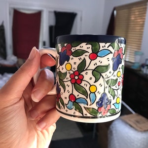 May include: A ceramic mug with a white handle and a floral design. The mug features a white base with a pattern of red, blue, and yellow flowers, green leaves, and a dark blue rim.