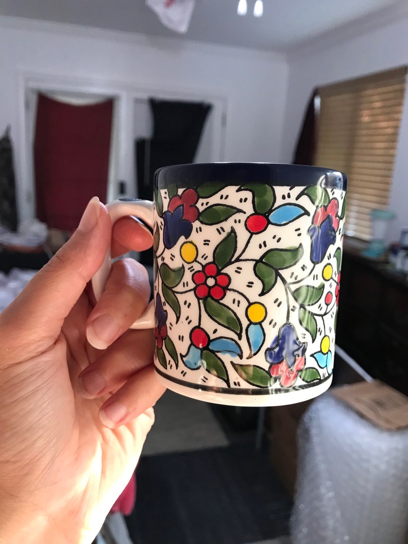 May include: A ceramic mug with a white handle and a floral design. The mug features a white base with a pattern of red, blue, and yellow flowers, green leaves, and a dark blue rim.
