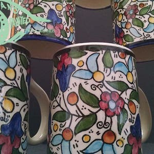 May include: A collection of ceramic mugs with a floral pattern. The mugs feature a white base with a colorful design of red, blue, and yellow flowers, green leaves, and orange accents. Each mug has a white handle and a blue rim.