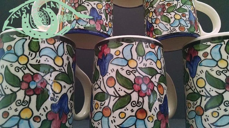 May include: A collection of ceramic mugs with a floral pattern. The mugs feature a white base with a colorful design of red, blue, and yellow flowers, green leaves, and orange accents. Each mug has a white handle and a blue rim.