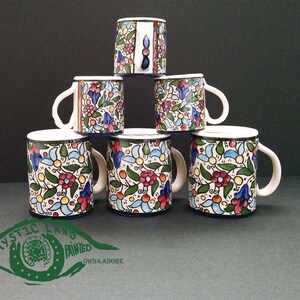 May include: Six ceramic mugs with white handles and rims, stacked in a pyramid. The mugs feature a colorful floral pattern with red, blue, and yellow flowers on a cream background. The base of each mug is dark blue.