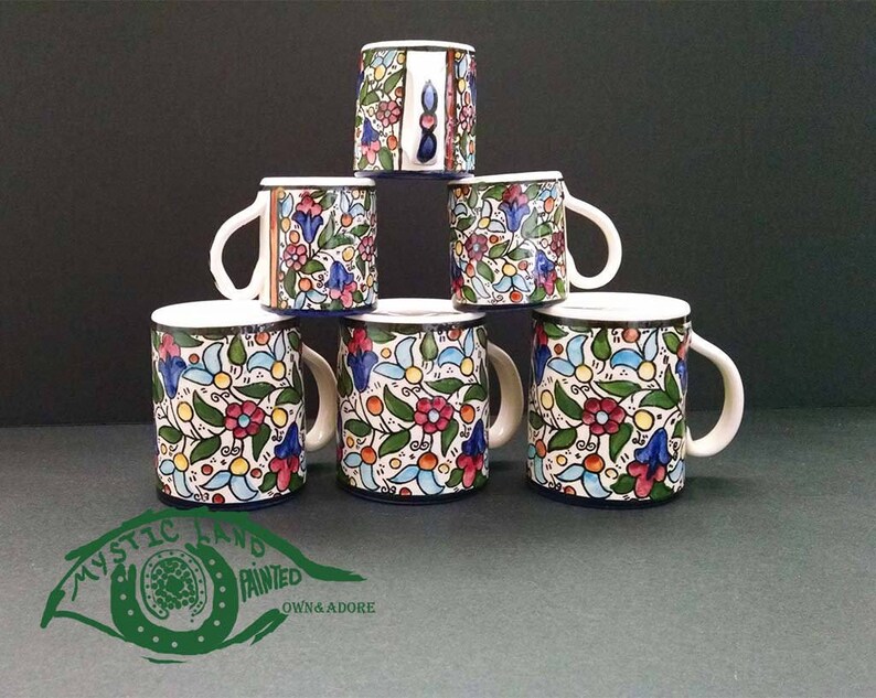 May include: Six ceramic mugs with white handles and rims, stacked in a pyramid. The mugs feature a colorful floral pattern with red, blue, and yellow flowers on a cream background. The base of each mug is dark blue.