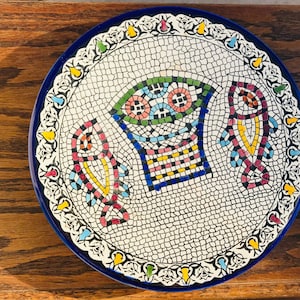 May include: A decorative ceramic plate with a mosaic design. The plate features a white background with a mosaic pattern and a blue rim. The mosaic design includes colorful fish and a basket.