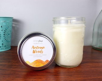 May include: A soy candle in a mason jar and a tin with a label that reads "Autumn Woods." The candle is white, and the label has orange and yellow watercolor accents. A teal metal container and a glass bottle are in the background.
