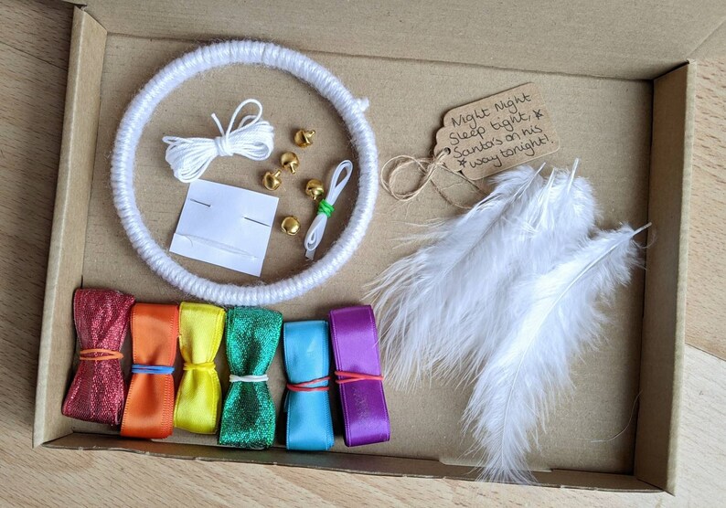 May include: A craft kit in a cardboard box, featuring a white yarn-wrapped hoop, white feathers, colorful ribbons, gold bells, and a tag that reads "Night Night Sleep tight, Santa's on his way tonight!"