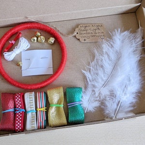 May include: A craft kit in a cardboard box, featuring a red wrapped ring, gold bells, white feathers, ribbons in red, gold, and green, and a tag with handwritten text. The kit includes various craft supplies for a DIY project.