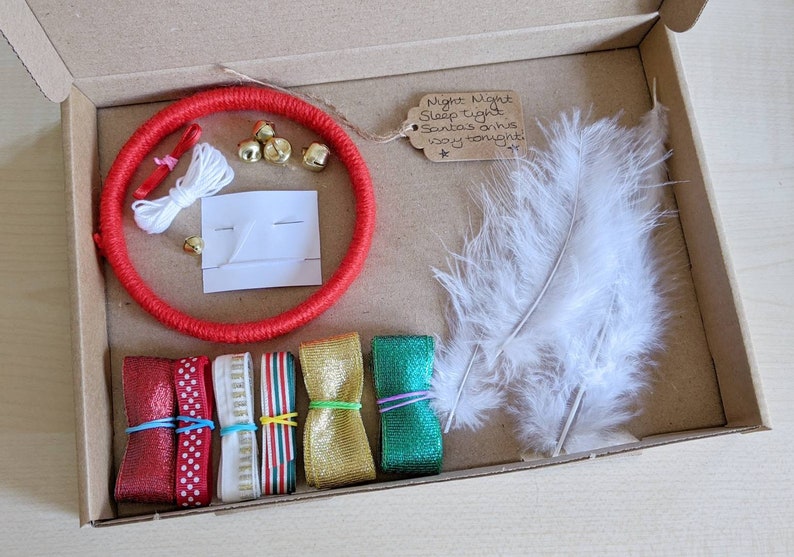 May include: A craft kit in a cardboard box, featuring a red wrapped ring, gold bells, white feathers, ribbons in red, gold, and green, and a tag with handwritten text. The kit includes various craft supplies for a DIY project.