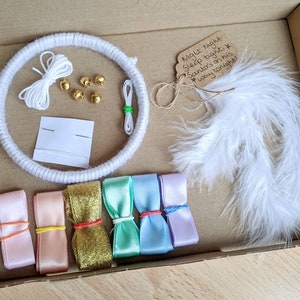 May include: A craft kit in a cardboard box, featuring a white yarn-wrapped hoop, white string, gold bells, needles, white feathers, and colorful ribbons. A tag reads "Night Night Sleep tight, Santa's on his way tonight!"