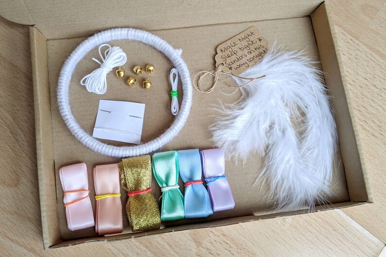 May include: A craft kit in a cardboard box, featuring a white yarn-wrapped hoop, white string, gold bells, needles, white feathers, and colorful ribbons. A tag reads "Night Night Sleep tight, Santa's on his way tonight!"