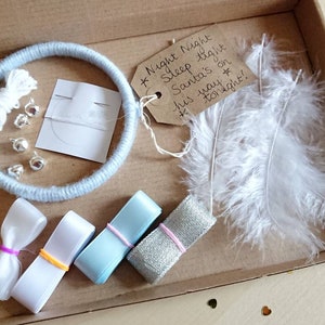 May include: A craft kit in a cardboard box, featuring a light blue yarn-wrapped ring, white feathers, ribbons, silver bells, and a tag with handwritten text. The tag reads "Night Night Sleep tight Santa's on his way tonight!"
