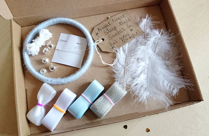 May include: A craft kit in a cardboard box, featuring a light blue yarn-wrapped ring, white feathers, ribbons, silver bells, and a tag with handwritten text. The tag reads "Night Night Sleep tight Santa's on his way tonight!"