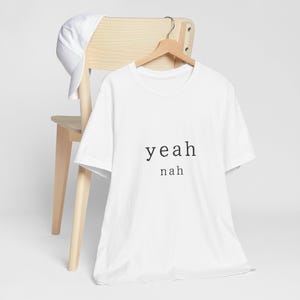 May include: A white short-sleeved t-shirt featuring the black text yeah nah printed on the front. The shirt is displayed on a wooden hanger, and a matching white baseball cap is draped over the top corner of the wooden chair back.