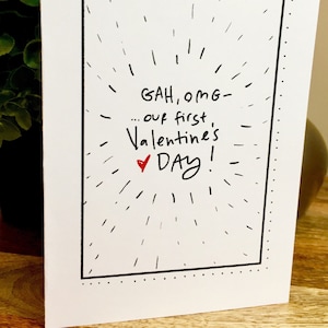 May include: A white card with black border and black text that reads "GAH, OMG - ... our first Valentine's DAY!" with a red heart symbol.