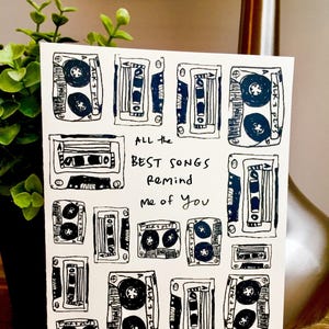 May include: A white card with a black and white illustration of cassette tapes. The text on the card reads "ALL the BEST SONGS remind me of you" and "I Lala Love You".