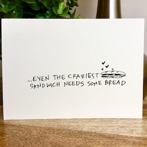 May include: A white card with black text that reads "...even the craziest sandwich needs some bread". A simple black and white drawing of a sandwich with a pickle and a tomato is above the text. There are three small hearts above the sandwich.