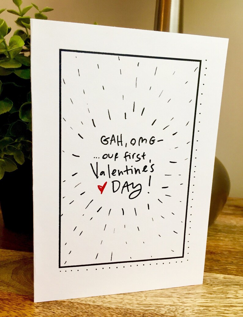 May include: A white card with black border and black text that reads "GAH, OMG - ... our first Valentine's DAY!" with a red heart symbol.