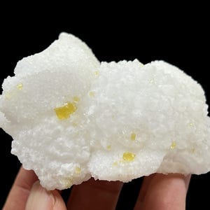 Peut inclure: Spécimen minéral blanc avec une surface texturée et cristalline. De petits cristaux jaune vif sont dispersés. Le minéral est posé sur un fond noir, soulignant sa couleur et sa texture.