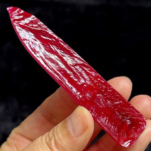 Può includere: Una gemma rossa vibrante e allungata con una superficie lucida e riflettente. La gemma è tenuta contro uno sfondo scuro, mostrando il suo colore ricco e la sua consistenza naturale. La forma è rettangolare con un'estremità appuntita.