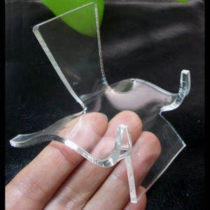 May include: A clear acrylic display stand for a small object, such as a crystal or a figurine. The stand has a curved base and a single upright support.