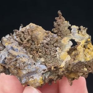Pu&ograve; includere: Un ammasso di cristalli di rame con una struttura dendritica ramificata. I cristalli sono una miscela di colori marrone, oro e giallo, con alcune aree di colore blu-grigio. I cristalli sono posizionati su uno sfondo scuro.