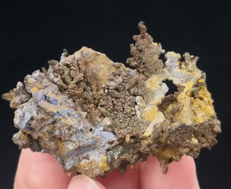 Pu&ograve; includere: Un ammasso di cristalli di rame con una struttura dendritica ramificata. I cristalli sono una miscela di colori marrone, oro e giallo, con alcune aree di colore blu-grigio. I cristalli sono posizionati su uno sfondo scuro.
