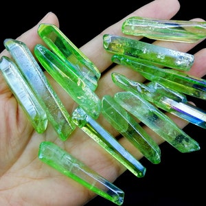 May include: A collection of green iridescent quartz points, also known as angel aura quartz, with a rainbow sheen. The points are various sizes and shapes, and are arranged on a hand.