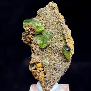 Pode incluir: Uma imagem de perto de um espécime mineral com vários cristais verdes vibrantes embutidos em uma matriz áspera e texturizada. A matriz tem tons marrons e dourados, com pequenas inclusões redondas laranja-amarelo. Os cristais são translúcidos e angulares.