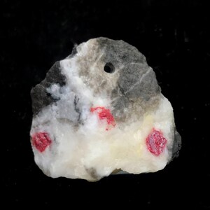 Op de afbeelding: Een ruwe steen met een gat erdoor. De steen heeft een mix van witte, grijze en zwarte kleuren, met drie heldere rode vlekken. Dit kan een mineraalmonster of een decoratief item zijn.