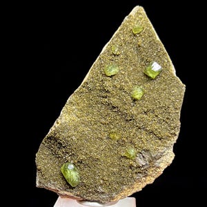 Pode incluir: Um espécime mineral natural com uma matriz áspera e texturizada em tons de ouro e castanho. Vários cristais verdes translúcidos e vibrantes estão embutidos na matriz, criando um contraste marcante. O espécime é exibido contra um fundo preto.