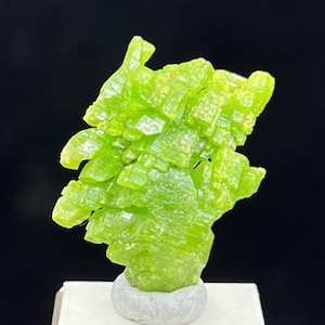 May include: A vibrant, lime-green mineral specimen with a crystalline structure. The mineral is mounted on a small, gray, rounded base, set against a dark background. The crystals have a bright, almost fluorescent appearance.