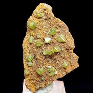 Pode incluir: Um espécime mineral bruto castanho com numerosos cristais pequenos, verde-brilhante, espalhados pela sua superfície. Os cristais têm uma forma geométrica e variam em tamanho. O mineral é exibido contra um fundo preto.