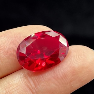May include: A faceted, oval-shaped, deep red gemstone. The gem is held between fingers against a black background. The stone's cut creates many reflective surfaces, enhancing its brilliance and colour.