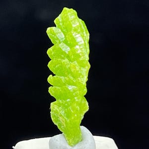 May include: A vibrant, lime-green mineral specimen with a crystalline structure. The mineral is elongated and features a layered, textured appearance. The background is black, highlighting the bright color and unique form of the mineral.