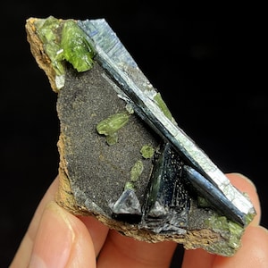 May include: A mineral specimen featuring a dark gray matrix with green and blue-gray crystal formations. The crystals have a prismatic shape and are partially embedded in the matrix. The specimen is held in a hand against a black background.