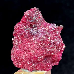 May include: A striking mineral specimen, predominantly magenta in colour, with a crystalline structure. The surface is covered in small, sparkling crystals. The mineral sits on a light-coloured base against a black backdrop.