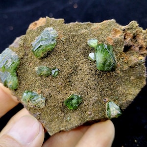 May include: A close-up of a rock with several green crystals embedded in it. The crystals are sharp and have a glassy appearance.